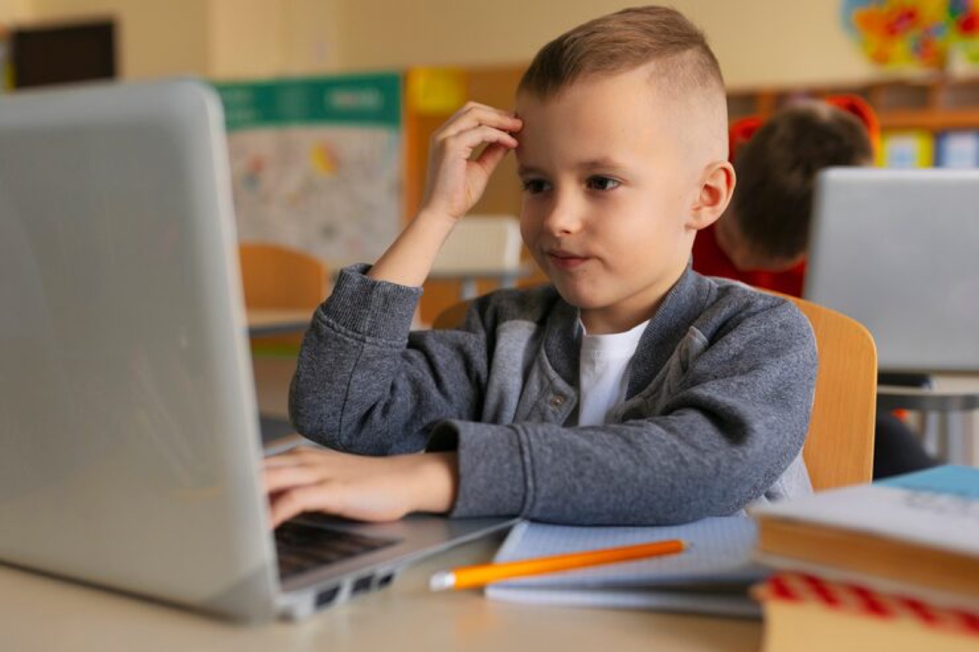 A boy pausing to think at his laptop in a classroom